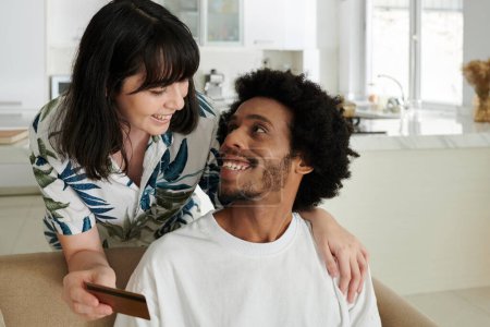 Téléchargez les photos : Heureux jeune homme afro-américain en t-shirt blanc assis sur le canapé et regardant sa femme avec carte de crédit lors de la discussion de la commande en ligne - en image libre de droit