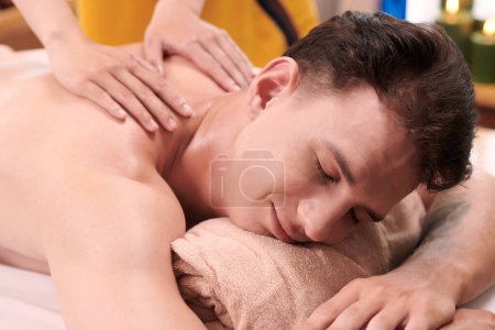 Foto de Joven hombre complacido acostado sobre su vientre en el sofá y recibiendo masaje de spa profesional de espalda y hombros por masajista trabajando en el salón - Imagen libre de derechos