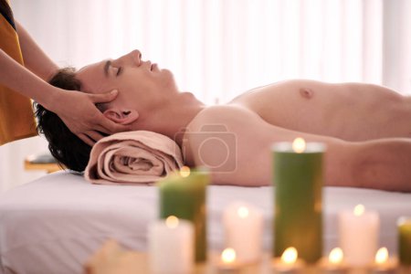 Photo for Side view of young serene and relaxed man lying on couch while masseuse standing behind and giving him professional massage in spa salon - Royalty Free Image
