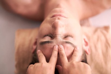 Téléchargez les photos : Au-dessus de l'angle du jeune homme serein se faire masser le visage dans un salon de spa luxueux tout en massant en appuyant sur son front pendant l'acupression - en image libre de droit