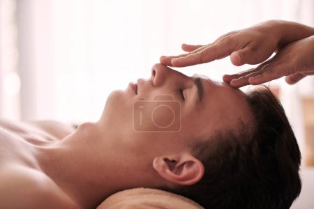 Photo for SIde view of young serene and relaxed man enjoying facial massage or acupressure while lying on couch or table in professional spa salon - Royalty Free Image