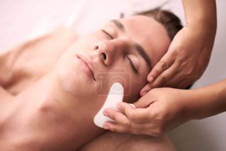 Téléchargez les photos : Jeune client masculin détendu de salon de spa obtenir luxueux gua sha massage facial bwing donné par masseuse professionnelle féminine - en image libre de droit