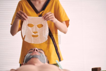 Foto de Joven esteticista femenina profesional en uniforme amarillo que sostiene la máscara de la hoja sobre el cliente masculino sereno con arcilla gris en la cara en el salón de spa - Imagen libre de derechos