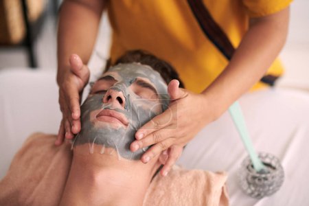 Photo for Focus on hands of young female beautician spreading hydrating sheet mask on face of relaxed male client with closed eyes visiting spa salon - Royalty Free Image