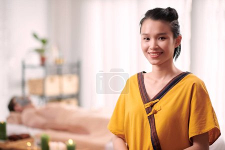 Foto de Joven mujer asiática sonriente esteticista y masajista en uniforme amarillo mirándote mientras está de pie frente a la cámara en el lujoso salón de spa - Imagen libre de derechos