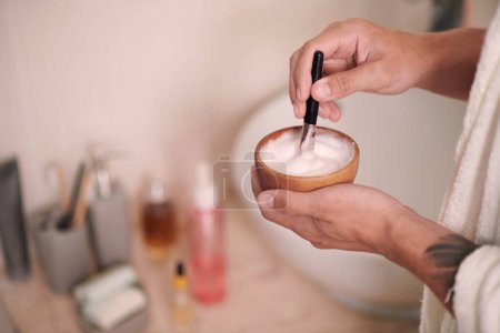Foto de Manos de hombre joven mezclando espuma suave con cepillo en un tazón de madera pequeño antes del procedimiento de afeitarse la cara en el baño por la mañana - Imagen libre de derechos