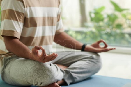 Téléchargez les photos : Gros plan d'un jeune homme actif en t-shirt et pantalon de survêtement assis sur le sol en pose de lotus tout en pratiquant le yoga à la maison - en image libre de droit