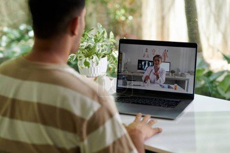 Téléchargez les photos : Focus sur l'écran d'ordinateur portable avec une jeune femme médecin en ligne en blouse de laboratoire assis par bureau dans un bureau médical et donnant une consultation au patient masculin - en image libre de droit
