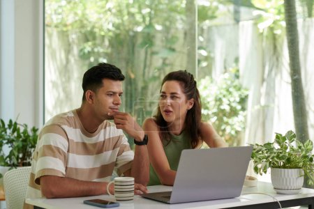Téléchargez les photos : Jeune couple interculturel assis par bureau devant un ordinateur portable et discuter point de travail tandis que l'homme sérieux en t-shirt rayé regardant l'écran - en image libre de droit