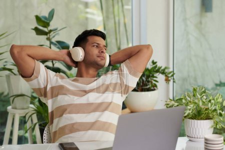 Téléchargez les photos : Jeune homme reposé dans les écouteurs et les vêtements décontractés regarder de côté par la fenêtre et écouter de la musique tout en étant assis devant l'écran de l'ordinateur portable - en image libre de droit
