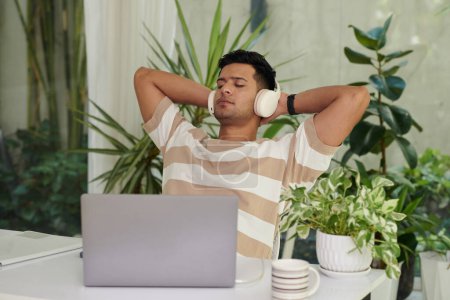 Foto de Joven hombre descansado en auriculares sentado junto al escritorio entre plantas verdes y escuchando música ambiental o sonidos de océano o lluvia en el descanso - Imagen libre de derechos