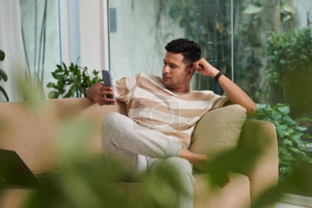 Photo for Young relaxed man sitting on beige comfortable couch and looking at smartphone screen while communicating in video chat with someone - Royalty Free Image