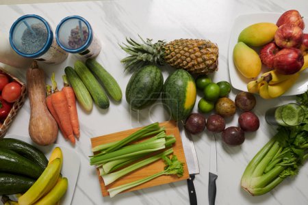 Photo for Top view of assortment of fresh, juicy and tasty fruits and vegetables surrounding wooden board with celery and two sharp knives on table - Royalty Free Image