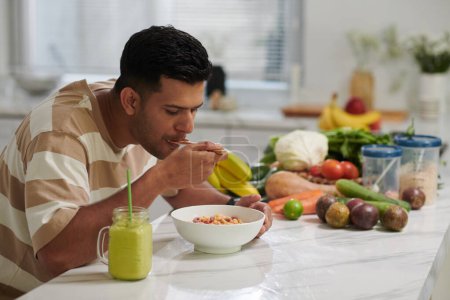 Foto de Hombre joven hambriento comiendo muesli del tazón y tomando jugo fresco o batido mientras está sentado junto a la mesa de la cocina con frutas y verduras - Imagen libre de derechos