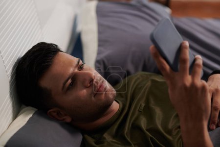 Foto de Hombre joven en camiseta mirando a través de las noticias en línea, viendo vídeo o charlando en el teléfono inteligente mientras está acostado en la cama por la mañana o por la noche - Imagen libre de derechos
