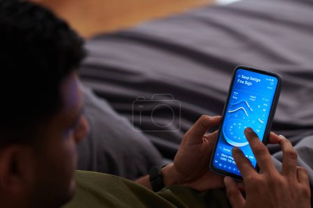 Photo for Focus on hands of young man choosing time settings in smartphone while lying on bed in the evening before sleep and switching on alarm - Royalty Free Image