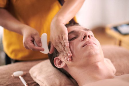 Téléchargez les photos : Esthéticienne massage visage de jeune homme avec gua sha outil - en image libre de droit