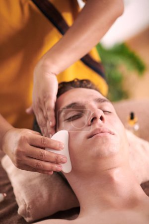Téléchargez les photos : Jeune homme obtenant gua sha massage du visage pour soulager la tension dans le visage et réduire les poches - en image libre de droit