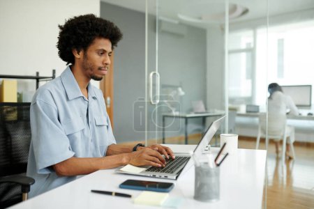 Téléchargez les photos : Sérieux jeune chef de projet noir travaillant sur ordinateur portable dans un bureau moderne - en image libre de droit