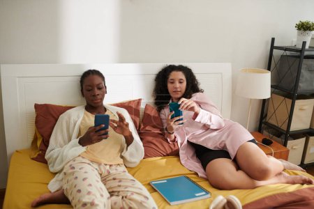 Foto de Chicas universitarias en pijama descansando en la cama y comprobando las notificaciones y mensajes en el teléfono inteligente - Imagen libre de derechos