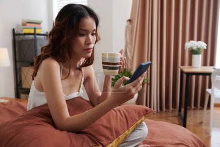 Téléchargez les photos : Jeune femme endormie assise au lit, buvant du café et faisant défiler les médias sociaux - en image libre de droit