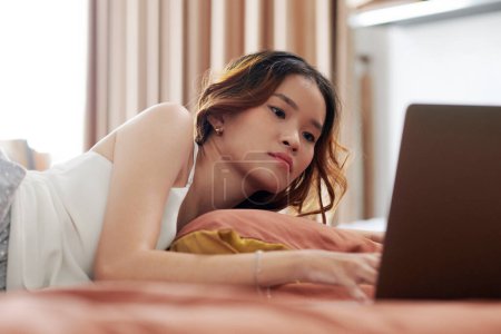 Téléchargez les photos : Jeune femme en pyjama regardant un film sur ordinateur portable quand elle est couchée sur le lit - en image libre de droit