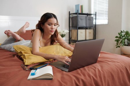 Foto de Estudiante universitaria acostada en la cama en su habitación y trabajando en el portátil - Imagen libre de derechos