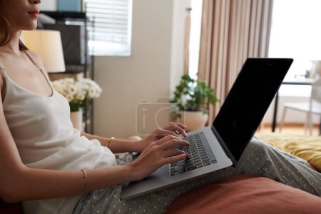 Téléchargez les photos : Image recadrée d'une jeune femme en tenue de salon travaillant sur un ordinateur portable au lit - en image libre de droit