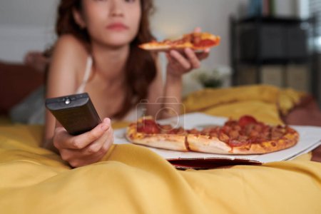 Téléchargez les photos : Image recadrée d'une jeune femme se relaxant sur son lit à la maison, mangeant de la pizza et regardant le spectacle à la télévision - en image libre de droit