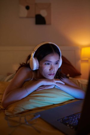 Téléchargez les photos : Fille dans les écouteurs regarder mélodrame sur ordinateur portable lorsqu'il est couché sur le lit - en image libre de droit