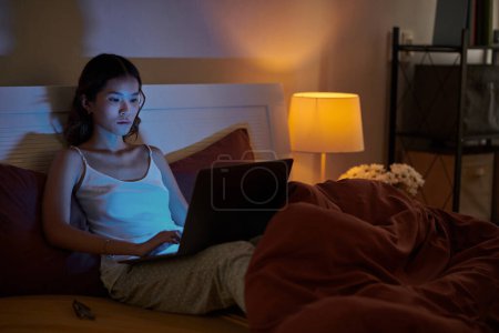 Foto de Mujer joven seria en pijama sentada en la cama y trabajando en la computadora portátil hasta tarde en la noche - Imagen libre de derechos