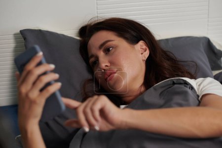 Téléchargez les photos : Jeune femme sérieuse couchée au lit et réglant l'alarme sur smartphone - en image libre de droit