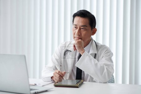Téléchargez les photos : Médecin généraliste pensif prenant des notes sur tablette lors d'un appel vidéo avec le patient - en image libre de droit