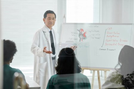 Téléchargez les photos : Médecin pointant vers le tableau blanc avec une carte cardiaque lorsque vous parlez à un groupe d'étudiants en médecine - en image libre de droit