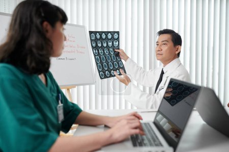Foto de Ortopedista analizando la imagen de los discos de la columna vertebral y pidiendo a la enfermera que llene el formulario en el portátil - Imagen libre de derechos