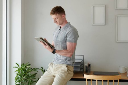 Téléchargez les photos : Jeune homme assis sur le bureau et lisant un document ou un article sur tablette - en image libre de droit