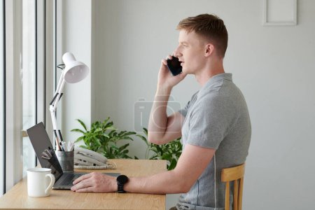 Photo for Young man sitting at desk in front of window and taking on phone - Royalty Free Image