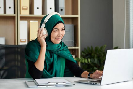 Téléchargez les photos : Sourire femme mûre en hijab travaillant sur ordinateur portable au bureau - en image libre de droit