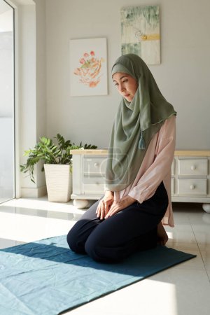 Téléchargez les photos : Femme d'âge moyen calme dans le hijab assis sur le tapis à la maison et priant - en image libre de droit