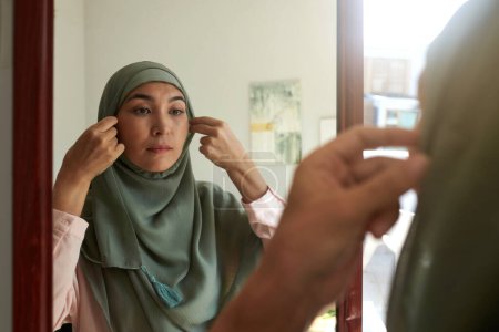 Foto de Mujer de mediana edad mirando al espejo y poniéndose hijab antes de salir del apartamento - Imagen libre de derechos