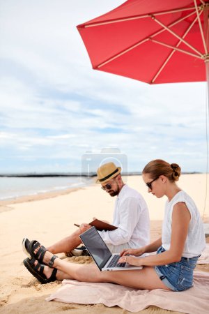 Téléchargez les photos : Nomades numériques assis sous le parapluie de plage, travaillant sur ordinateur portable et livre de lecture - en image libre de droit