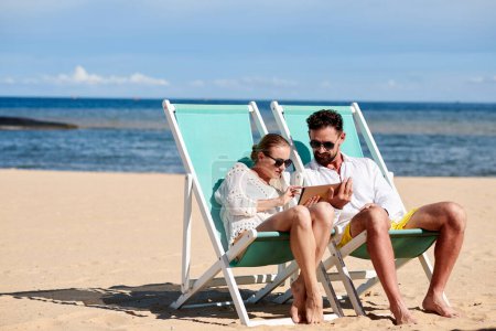 Foto de Pareja en gafas de sol sentados en chaise-lounges en la playa soleada y reservar city tour - Imagen libre de derechos