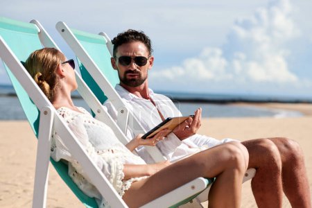 Téléchargez les photos : Homme fronçant les sourcils parler à petite amie quand ils sont bronzer dans des chaises longues sur la plage - en image libre de droit
