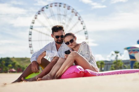 Téléchargez les photos : Couple souriant en lunettes de soleil assis sur des matelas flottants et vérifier les nouvelles sur les médias sociaux, ferris roue en arrière-plan - en image libre de droit