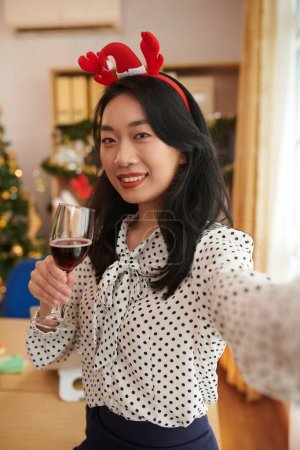 Foto de Retrato de mujer emprendedora alegre sosteniendo copa de vino al tomar selfie en la oficina decorado para Navidad - Imagen libre de derechos
