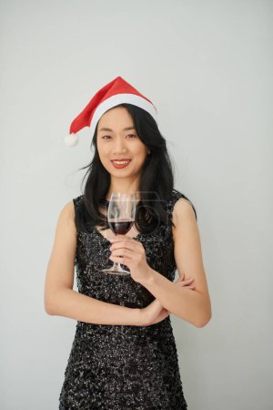 Téléchargez les photos : Portrait de jeune femme souriante en robe d'étincelle et chapeau santa claus tenant un verre de vin rouge - en image libre de droit