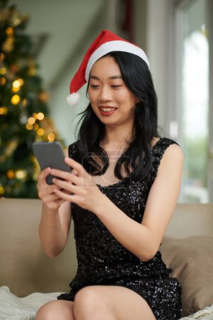 Photo for Portrait of smiling young woman sending Christmas greetings to friends via messenger mobile app - Royalty Free Image