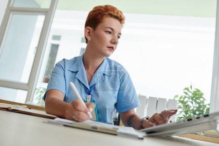Foto de Enfermera médica seria verificando datos en computadora y tomando notas en documento - Imagen libre de derechos