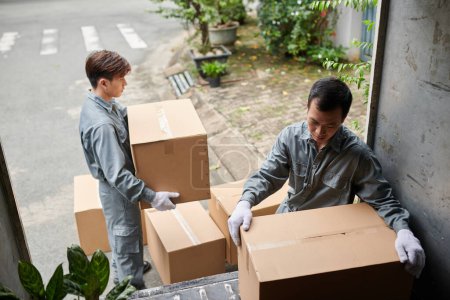 Photo for Movers in grey uniform loading cardboard boxes with belongings on client in van - Royalty Free Image