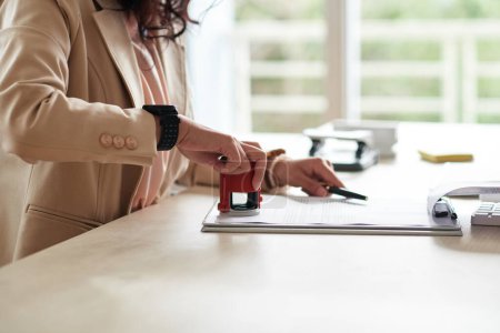Téléchargez les photos : Gros plan de la jeune femme d'affaires apposant son timbre sur un document à table au bureau - en image libre de droit